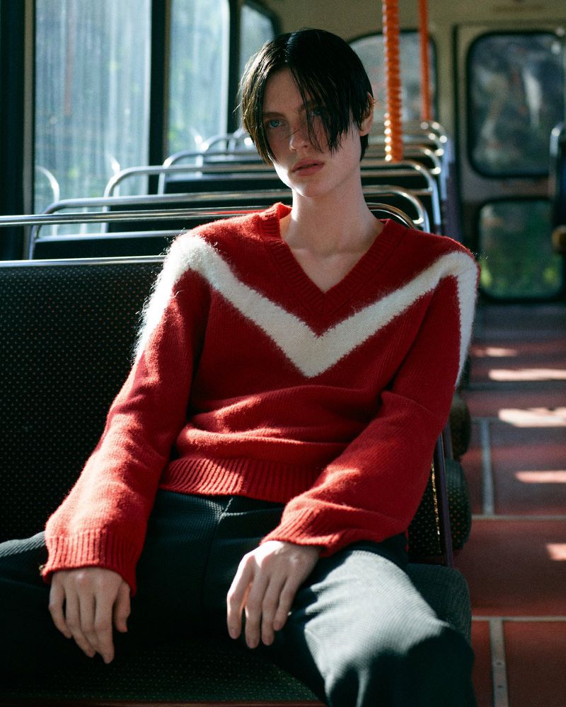 Portrait of a young adult male with dark hair wearing a red and white sweater, sitting in what appears to be a bus interior.