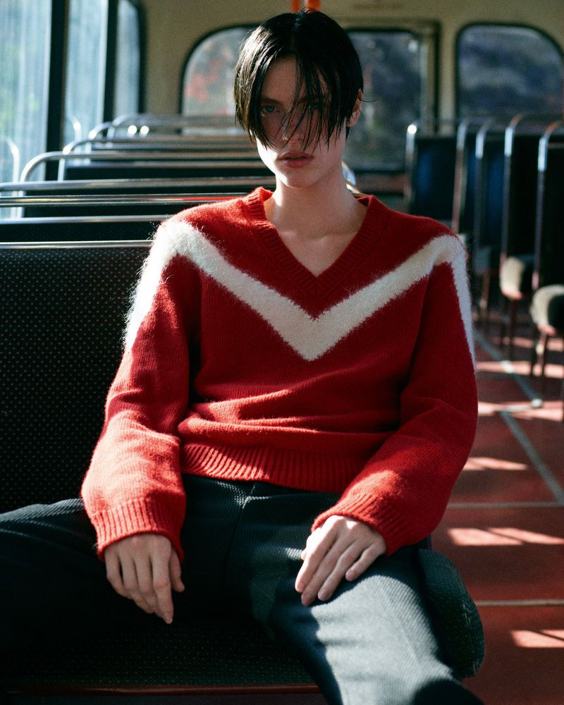 A young adult poses on a bus in a red and white sweater, with dark hair framing their face, creating a portrait-style image with a vintage feel.