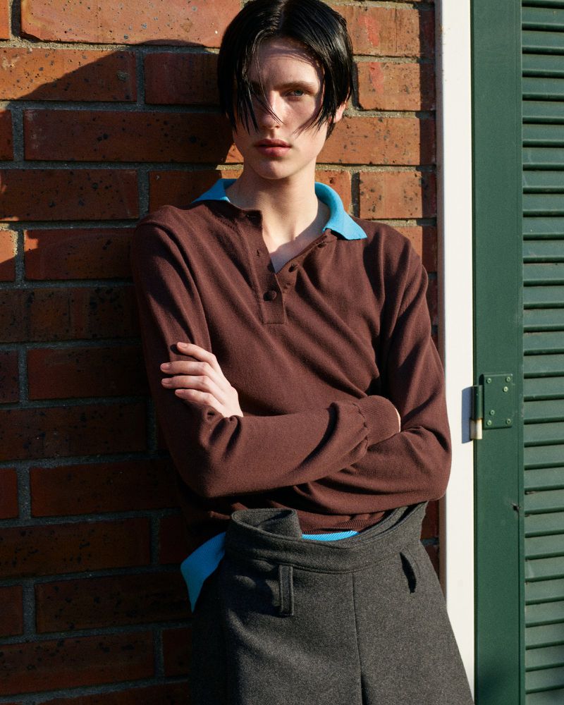 A young adult woman in brown top with blue collar and grey pants stands against a brick wall and green shutters, facing the camera.