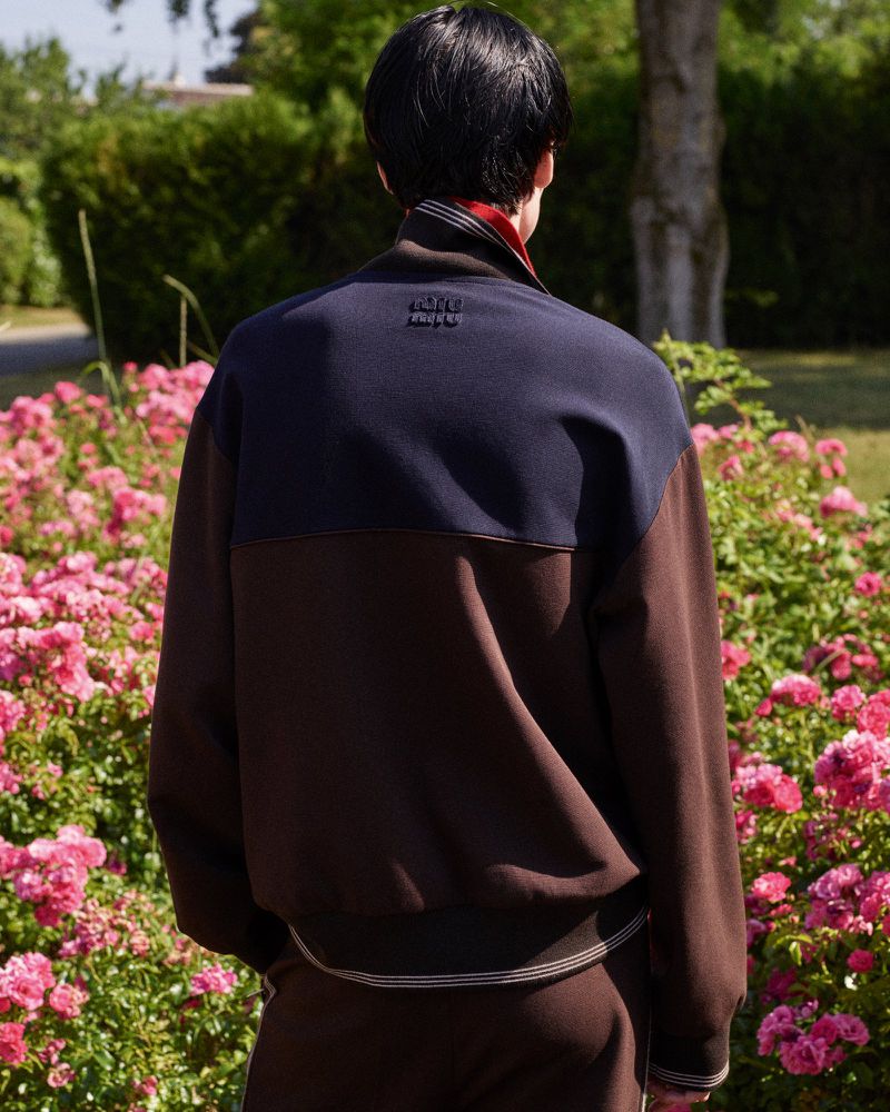 Back view of a person in a color-blocked brown and navy sweater standing in a garden with pink roses on a sunny day.