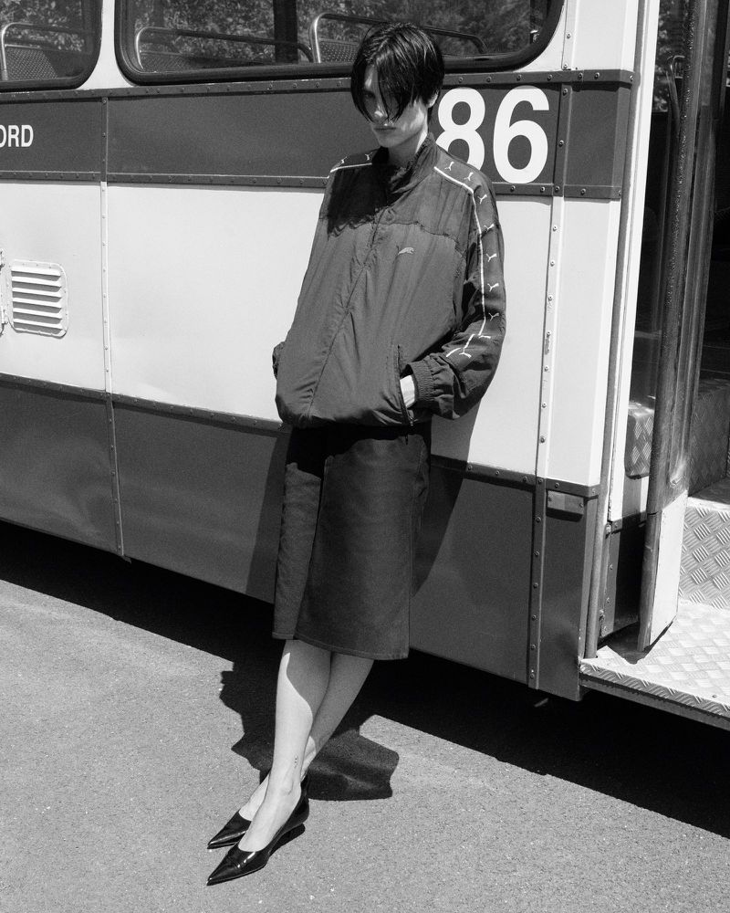 Woman in red windbreaker and skirt posing against bus in a black and white outdoor fashion shot with natural light.