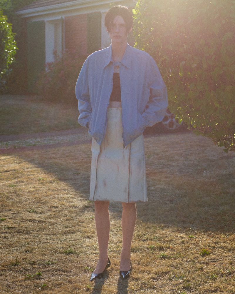 Elegant woman stands in yard with oversized bomber jacket and neutral skirt, accessorized with pointed-toe heels, illuminated by sunlight.