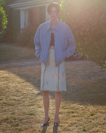 Elegant woman stands in yard with oversized bomber jacket and neutral skirt, accessorized with pointed-toe heels, illuminated by sunlight.
