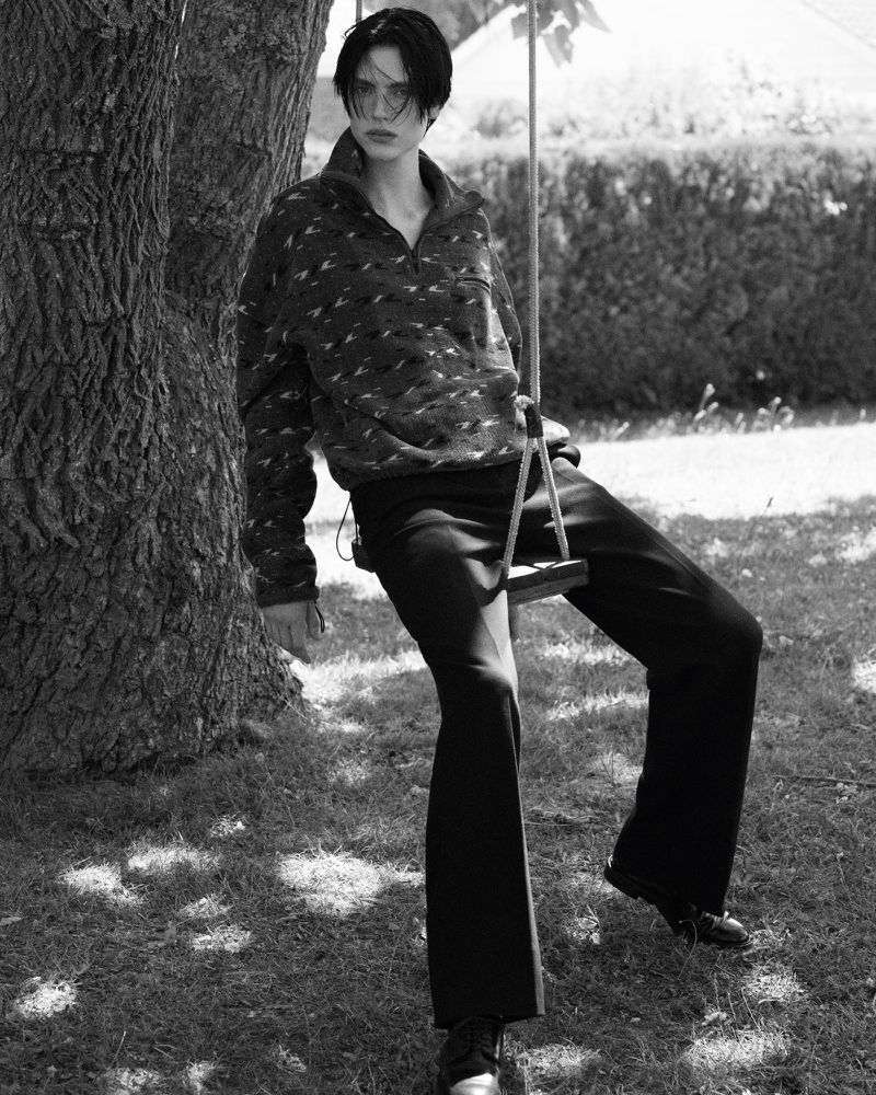 Black and white portrait of a young adult wearing patterned pullover and dress pants sitting on a wooden swing in a grassy park.