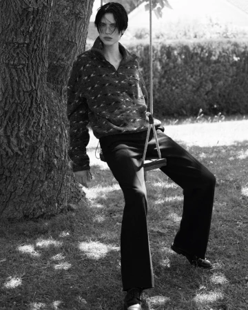 Black and white portrait of a young adult wearing patterned pullover and dress pants sitting on a wooden swing in a grassy park.