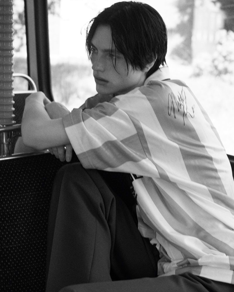 Monochrome medium shot of a young adult male model with dark hair sitting indoors wearing a striped shirt and dark pants.