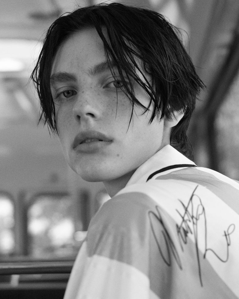 Monochrome portrait of a young adult male with dark hair, gazing intently at the camera, wearing a striped shirt with signature on back.