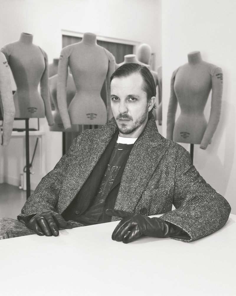 Portrait of a man in tweed coat with leather gloves sitting at a white table, with dress forms in the background, in black and white.