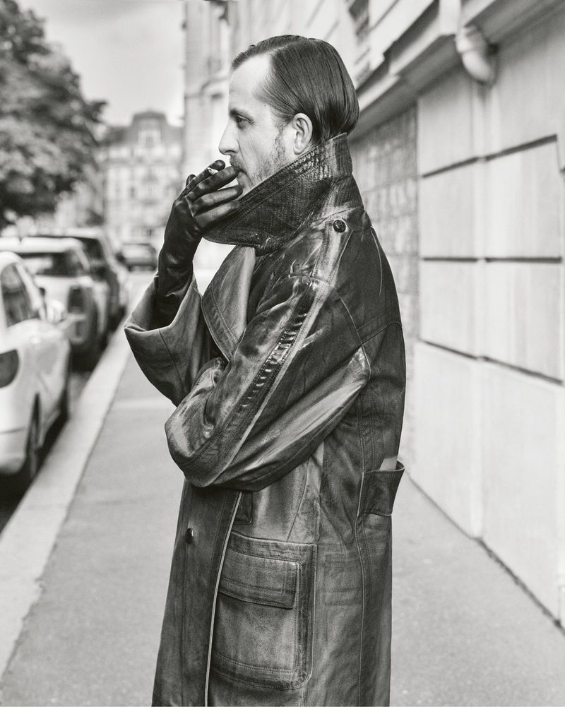 Black and white portrait of an adult male in profile, wearing a leather coat and gloves, standing on a city sidewalk near parked cars.
