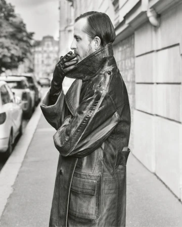 Black and white portrait of an adult male in profile, wearing a leather coat and gloves, standing on a city sidewalk near parked cars.