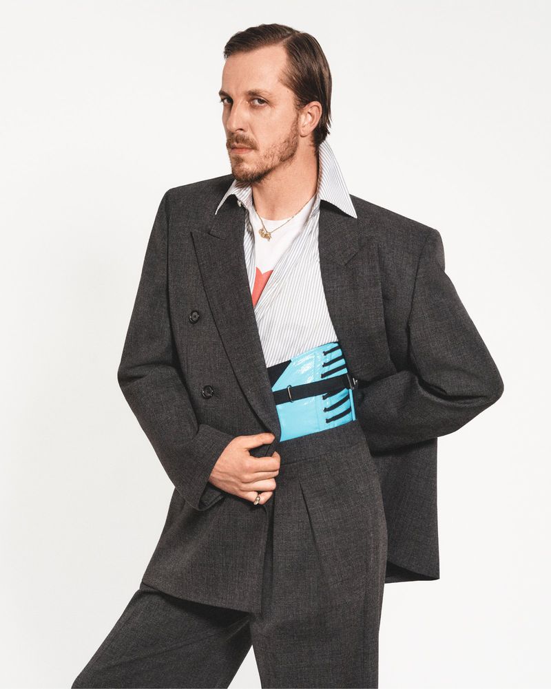 Man in a gray suit and striped shirt strikes a pose against a white backdrop. He is accessorized with a blue waist bag and gold jewelry.