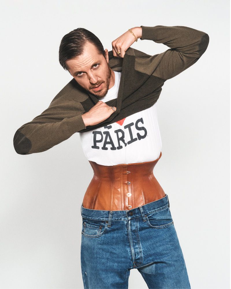Adult male in jeans and brown corset removing a crewneck sweater to reveal a white 'I love Paris' t-shirt in a bright studio setting.