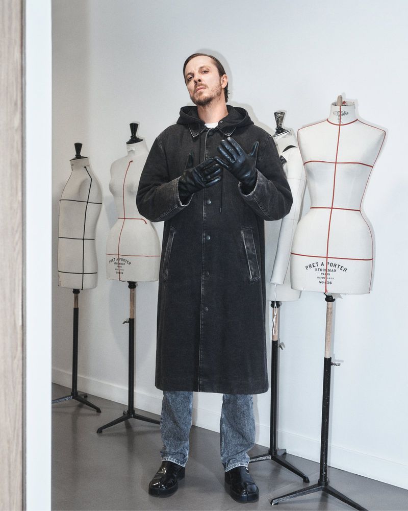 Man posing in a black coat and gloves in front of mannequin torsos in a minimalist studio, fashion, clothing, and style.