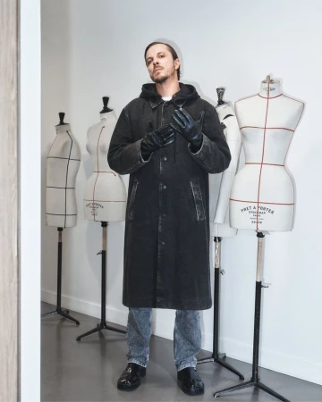 Man posing in a black coat and gloves in front of mannequin torsos in a minimalist studio, fashion, clothing, and style.