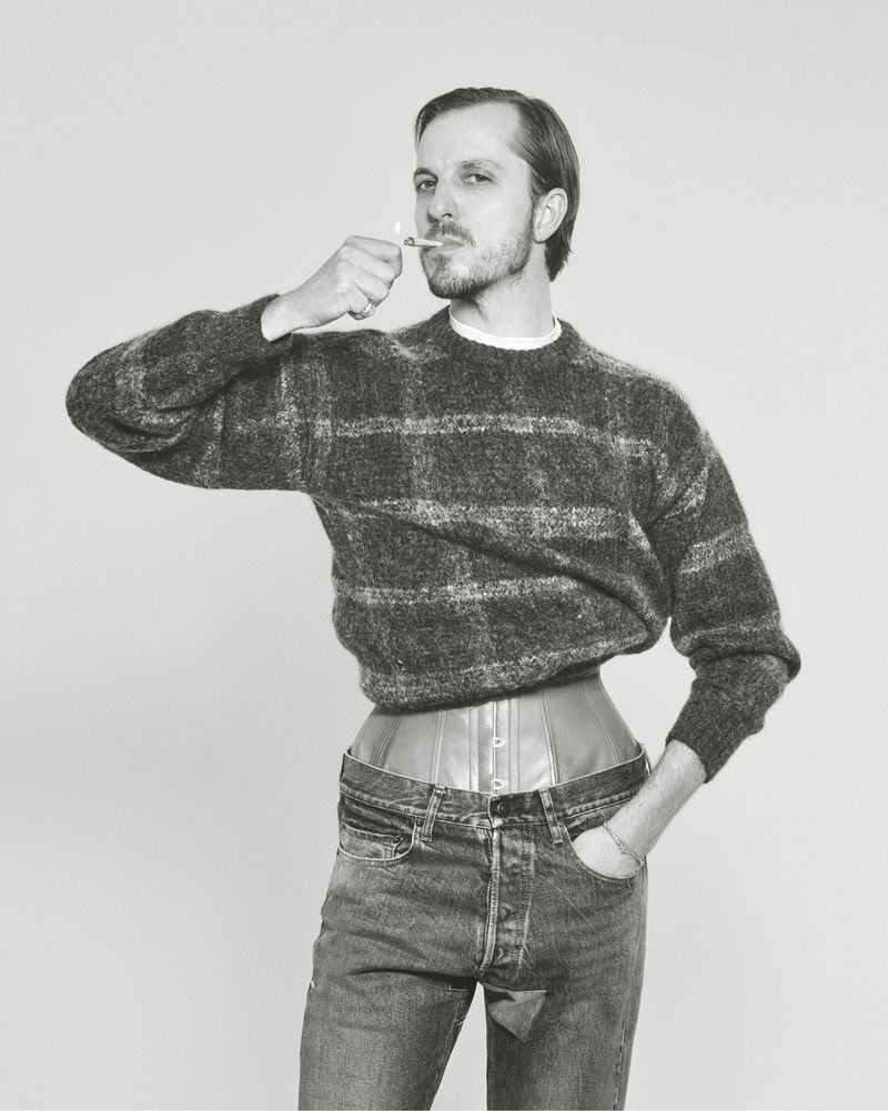 Monochrome studio shot of a man wearing a sweater, corset, and jeans while lighting a cigarette, set against a neutral background.
