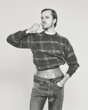 Monochrome studio shot of a man wearing a sweater, corset, and jeans while lighting a cigarette, set against a neutral background.