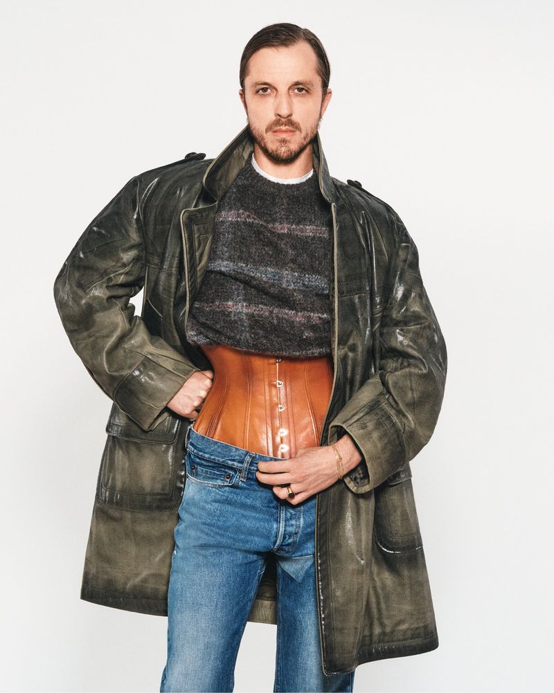 Man in a grey sweater and blue jeans wearing a brown leather corset and oversized green trench coat in a bright photo studio.