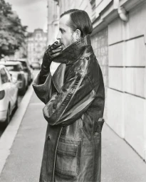 Black and white profile of Glenn Martens smoking a cigarette on a city sidewalk while wearing an oversized leather coat and gloves.