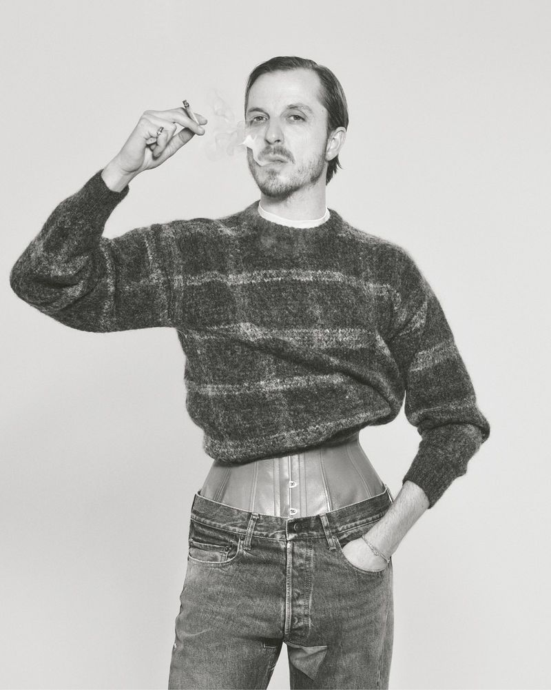 Glenn Martens in a patterned knit sweater, cinched corset, and jeans holding a smoking cigarette against a studio background.