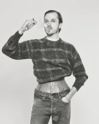 Glenn Martens in a patterned knit sweater, cinched corset, and jeans holding a smoking cigarette against a studio background.