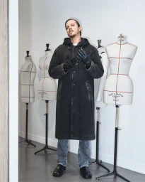 Man wearing a dark hooded coat and black leather gloves standing in a room with dressmakers mannequins.