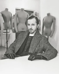 Glenn Martens seated at a white table wearing a herringbone coat and black leather gloves in front of tailor mannequins.