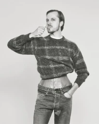Man wearing a plaid sweater and jeans lighting a cigarette against a plain light studio background
