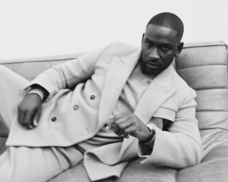 Black man in a light-colored suit reclines on a couch in a studio setting, looking at the camera with a serious expression. Monochromatic fashion shot.