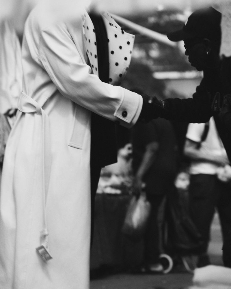 Two people in monochrome exchanging a handshake. One is wearing a light overcoat and polka dot hat; the other, a baseball cap.