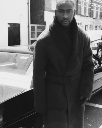 Monochrome portrait of an adult male with short hair, wearing a belted overcoat and standing next to a parked car on a city street.