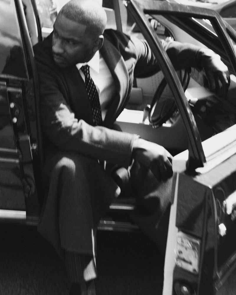 Monochrome portrait of a sharply dressed African-American man stepping out of a car, looking sideways with a serious expression.