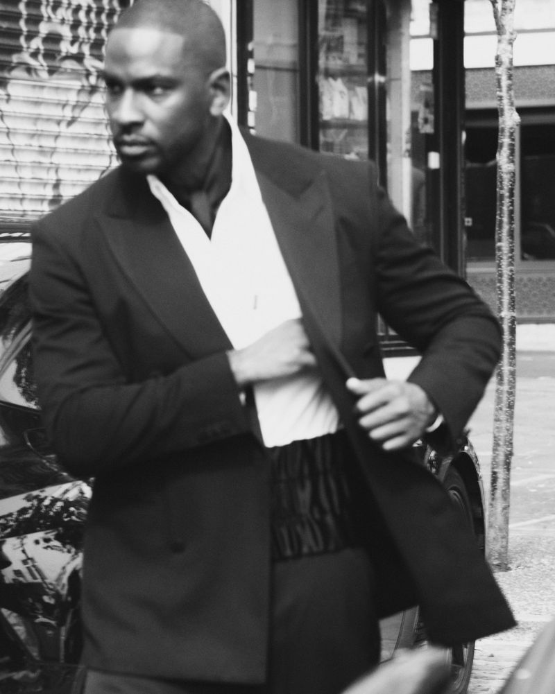Monochrome portrait of an adult male in a tuxedo with an open white dress shirt and black cummerbund, set in an urban environment.