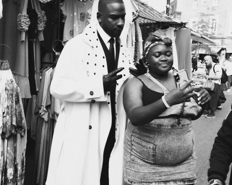Black and white shot of two people at an outdoor market: a man in a polka dot coat and a woman taking a picture with her phone.