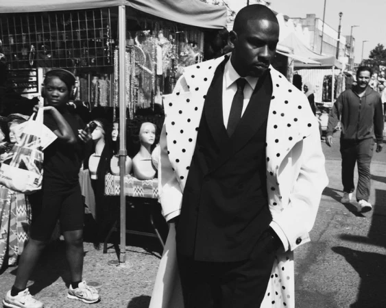 A black and white street scene captures a man in a polka dot coat walking past market stalls and pedestrians on a sunny day.