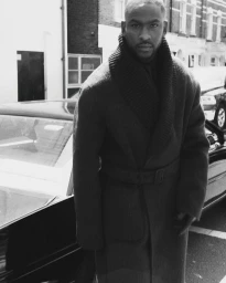 Skepta wearing a belted coat standing outdoors by a parked car for a Fantastic Man editorial portrait