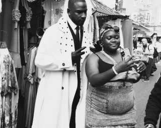 Black and white portrait of Skepta in a white coat posing with a fan taking a selfie at a market.