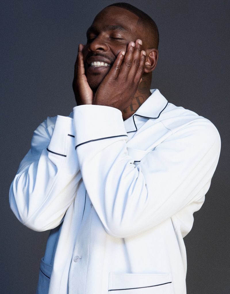 Skepta wearing white pyjamas with dark piping holding his face with both hands against a grey background for Fantastic Man