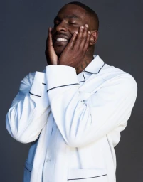 Skepta wearing white pyjamas with dark piping holding his face with both hands against a grey background for Fantastic Man