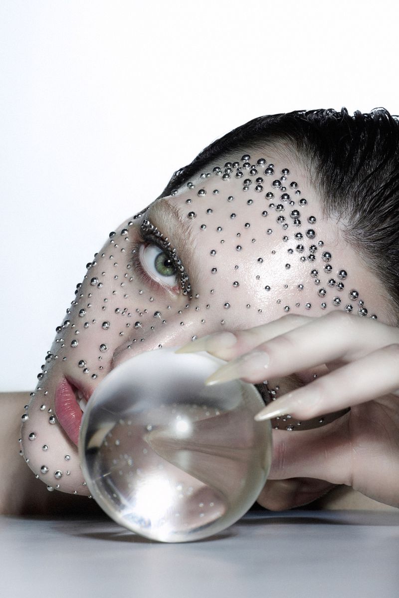 Close-up of a woman's face adorned with water droplets, featuring dramatic makeup, long nails, and holding a transparent crystal ball.