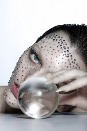 Close-up of a woman's face adorned with water droplets, featuring dramatic makeup, long nails, and holding a transparent crystal ball.