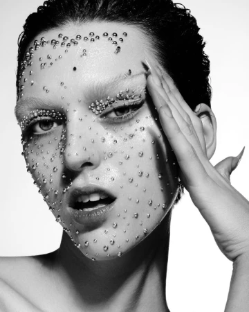 Black and white portrait of a woman with water droplets on her face and stylized eyebrows, touching her face with manicured nails.