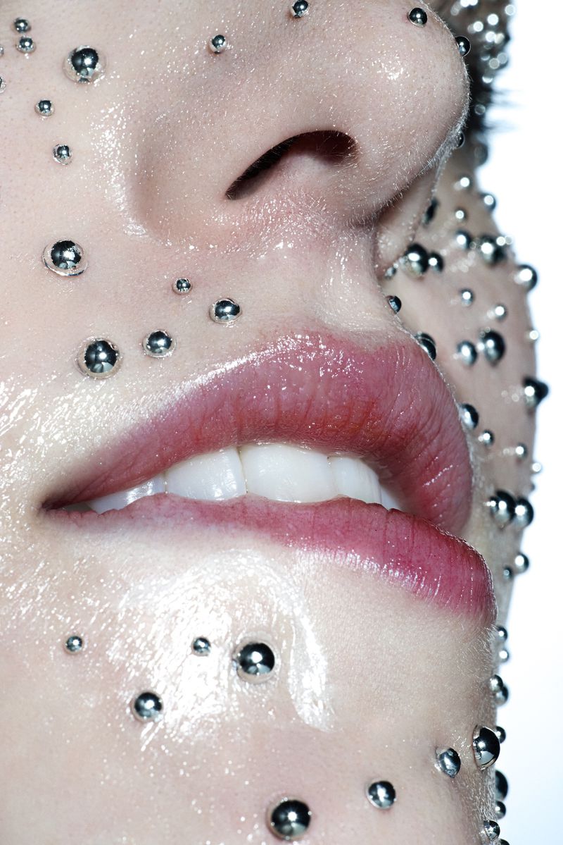 Close-up of a woman's face with a shiny, wet complexion, adorned with small, reflective silver spheres, highlighted against white background.