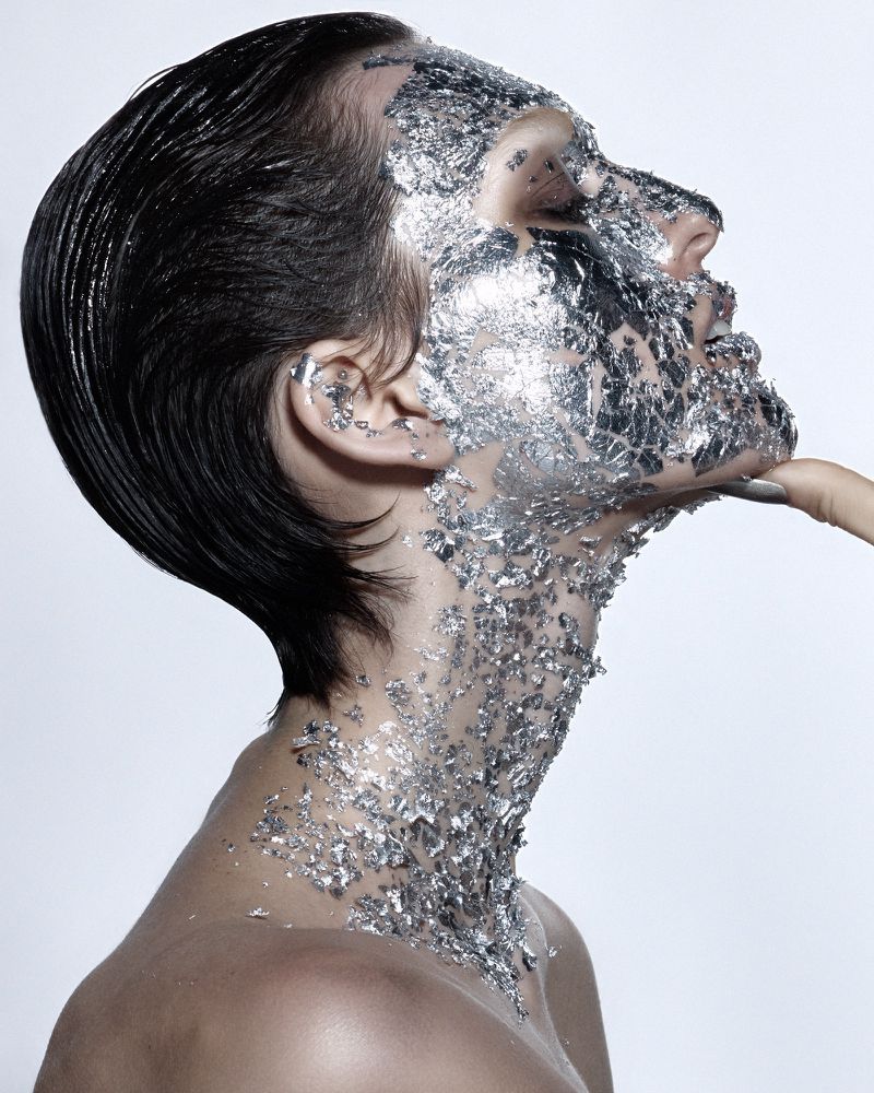 Adult female model with short dark hair and silver foil pieces adhering to her face and neck poses on a solid light background.