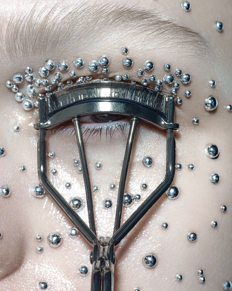 Close-up shot of an eye and eyelash curler surrounded by silver liquid beads on the skin, creating a surreal, beauty-focused image.