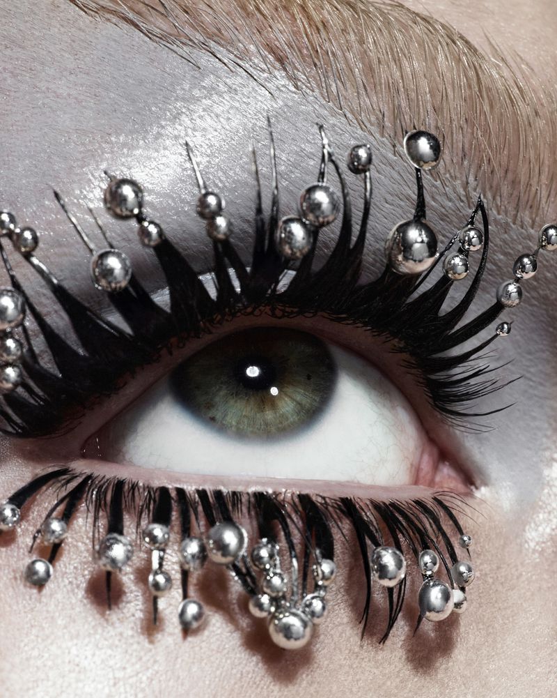 Close-up shot of a woman's eye enhanced with creative black eyelashes adorned with small silver spheres, showcasing avant-garde beauty.