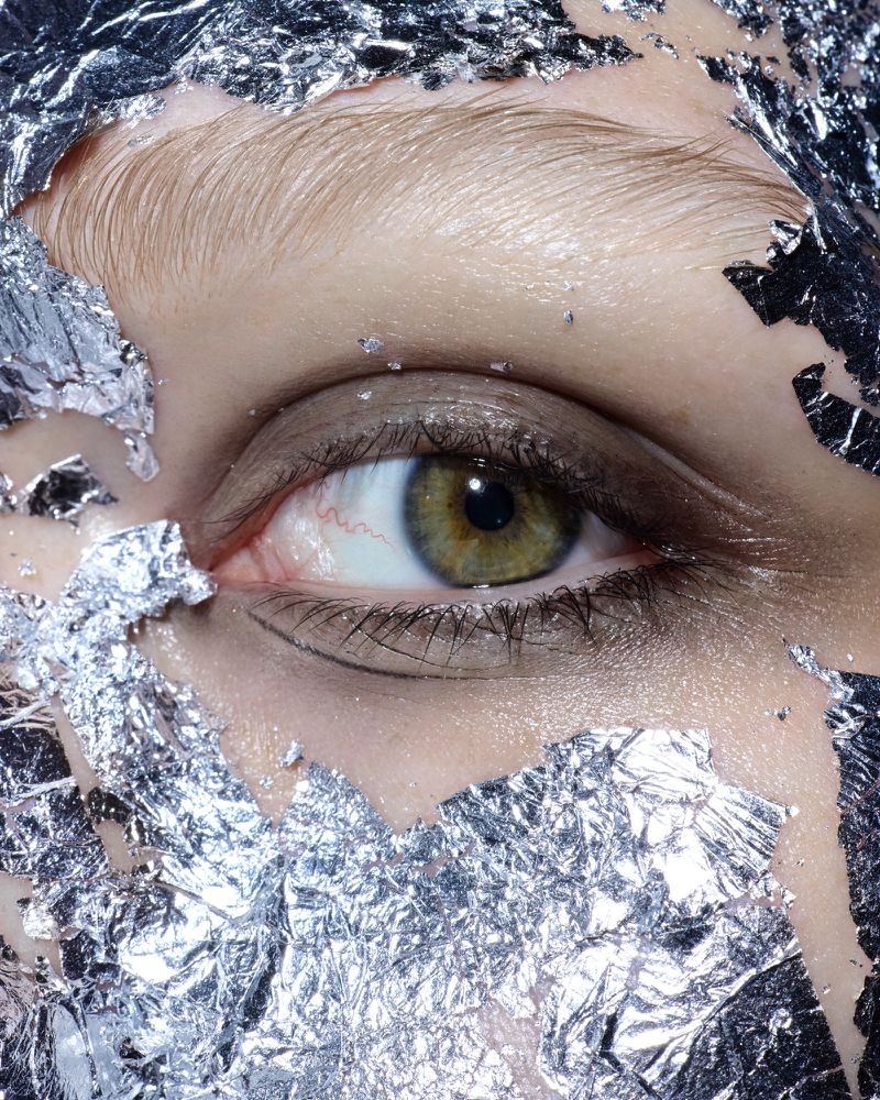 Close-up of a woman's eye surrounded by shiny silver foil. The focus is on the hazel eye with visible blood vessels.