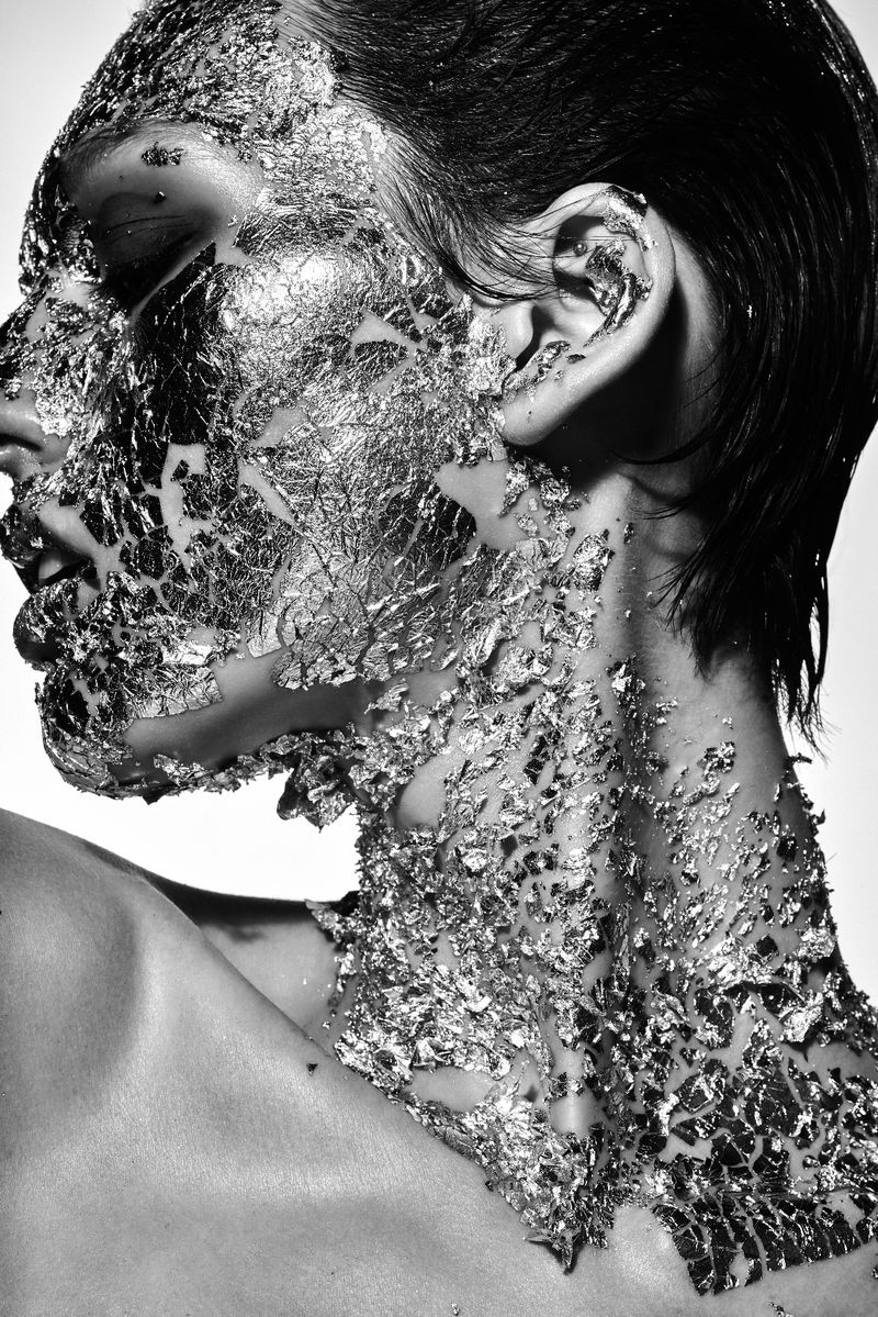 Black and white portrait of a woman with silver foil on her face and neck, eyes closed, against a bright white background.