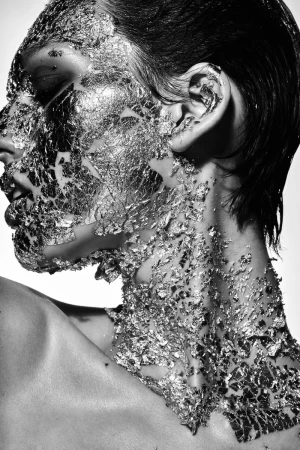 Black and white portrait of a woman with silver foil on her face and neck, eyes closed, against a bright white background.