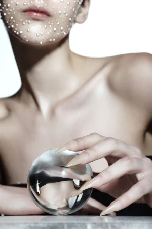 Close-up portrait of a woman with rhinestones on her face, holding a clear glass ball, set against a bright white backdrop.