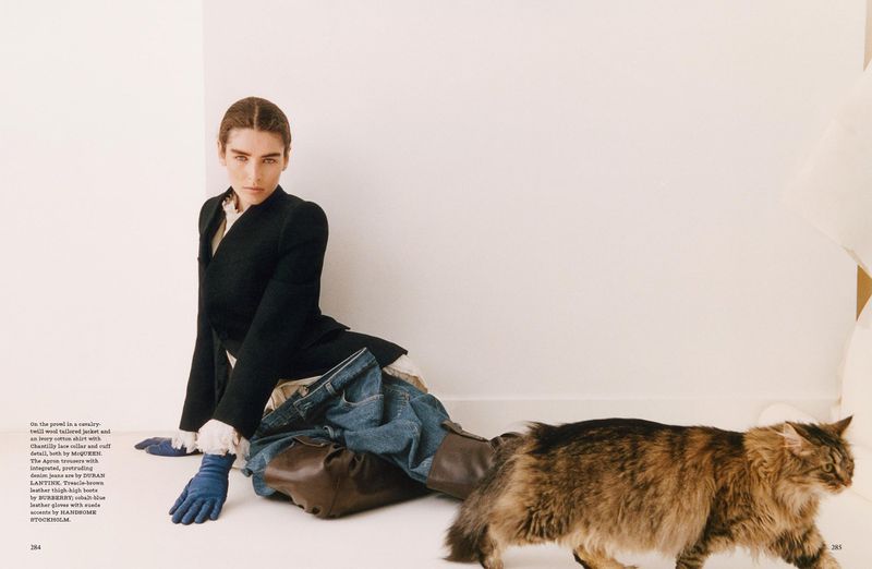 Woman poses with a brown long-haired cat in a fashion editorial shot. She wears a black blazer, denim, and blue gloves indoors.